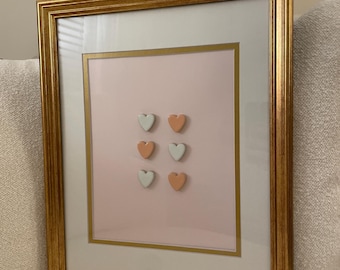 May include: A framed artwork featuring six heart-shaped objects in a gold frame. The hearts are arranged in two rows of three, with alternating colors of white and peach against a pink background. The frame has a gold border.