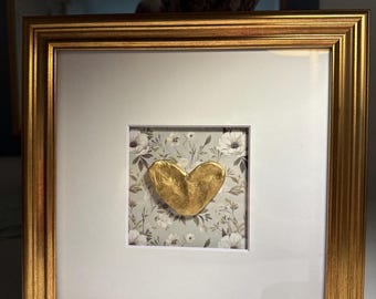 May include: A framed artwork featuring a gold-colored heart on a floral patterned background. The heart is centered within a white mat, encased in a gold-colored wooden frame. The artwork is a decorative piece, suitable for home decor.