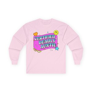 Könnte beinhalten: Hellrosa Langarmshirt mit dem Text "CERTIFIED DEADLY AUNTIE" in einem Retro-Schriftstil. Das Design beinhaltet ein Herz, ein Smiley, Sterne und eine Sonnenbrille. Es ist ein lässiges Kleidungsstück.