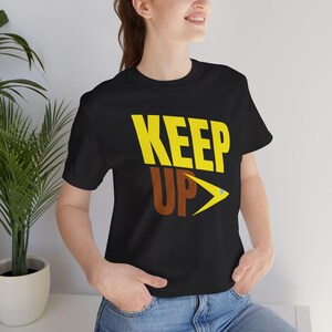 May include: Black t-shirt featuring the words "KEEP UP" in yellow and brown block letters. A yellow and blue arrow points to the right. The shirt is made of a soft material.
