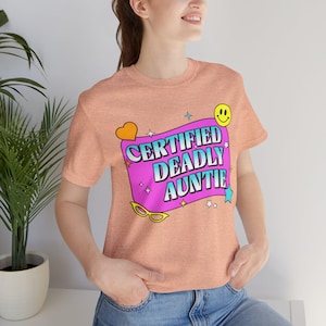 May include: Peach-colored t-shirt with the text "CERTIFIED DEADLY AUNTIE" in a colorful design. The graphic includes a pink rectangle, a yellow smiley face, and other decorative elements. The shirt is made of a soft material.