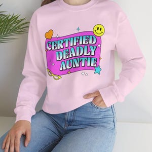 Könnte beinhalten: Hellrosa Sweatshirt mit dem Text "CERTIFIED DEADLY AUNTIE" in einem farbenfrohen Design. Die Grafik enthält ein Smiley, Sterne und ein Herz. Das langärmlige Oberteil wird mit einer blauen Jeans kombiniert.