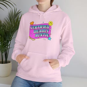 Peut inclure: Un sweat à capuche rose clair avec une poche avant et une capuche à cordon. Le devant présente un graphisme avec l'inscription "CERTIFIED DEADLY AUNTIE" dans un style rétro, avec un cœur, un smiley, une étoile et des lunettes.