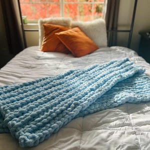 May include: A light blue chunky knit throw blanket on a white bed with orange and white throw pillows.