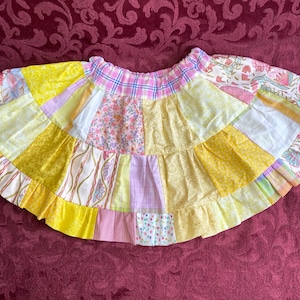 Puede incluir: Una falda de patchwork amarilla y rosa con un dobladillo con volantes. La falda está hecha de diferentes telas con patrones florales y geométricos.