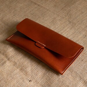 May include: A brown leather clutch with a curved flap and a snap closure. The edges are stitched with white thread. The clutch is rectangular and appears to be empty.