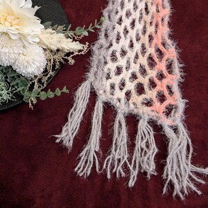 May include: A handmade, open-weave scarf with a gradient of colors, including gray, white, and peach. The scarf has a fringed edge and a soft, textured appearance. The scarf is likely made of a fuzzy yarn, and is a fashion accessory.