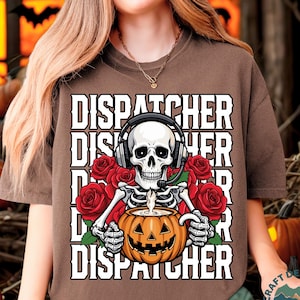 May include: Brown t-shirt with a Halloween-themed graphic. The design features a skeleton wearing a headset, holding a jack-o'-lantern, and surrounded by red roses. The word "DISPATCHER" is printed multiple times.