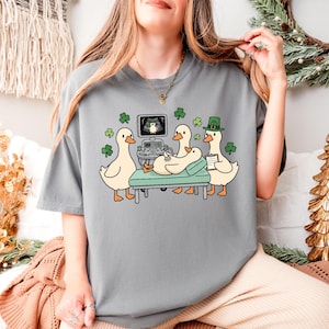 Könnte beinhalten: Graues T-Shirt mit einer Cartoon-Illustration von Enten in einer medizinischen Umgebung. Eine Ente liegt auf einem Behandlungstisch, während andere beobachten. Das Design enthält Kleeblätter und eine Ente mit grünem Hut. Der Ultraschallbildschirm ist sichtbar.