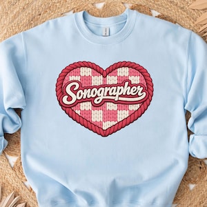 Könnte beinhalten: Hellblaues Sweatshirt mit herzförmigem Design. Das Herz hat einen roten Seilrand und ein rosa-weißes Karomuster mit dem Wort "Sonographer" in Schreibschrift.
