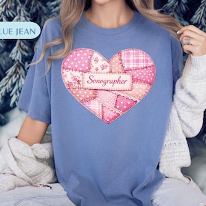 Könnte beinhalten: Ein blaues T-Shirt mit einem Patchwork-Herz-Design. Das Herz besteht aus verschiedenen rosa Stoffen, darunter Blumen- und Tupfenmuster. Das Wort "Sonographer" ist auf einem kleinen Etikett innerhalb des Herzens aufgedruckt.