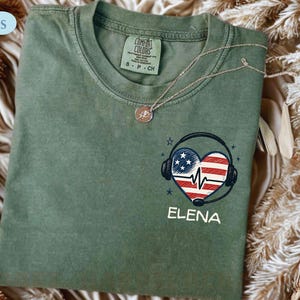 May include: An olive green t-shirt with a heart-shaped design featuring the American flag and headphones. The name "ELENA" is printed below the design. A silver necklace with a pendant rests on the shirt.