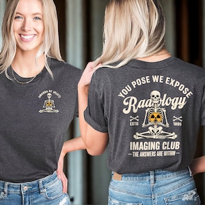 May include: Dark grey t-shirt featuring a skeleton graphic. The front of the shirt has the text "AMANDA RT (R)(CT)". The back of the shirt has the text "YOU POSE WE EXPOSE Radiology IMAGING CLUB - THE ANSWERS ARE WITHIN - ESTO 1895".