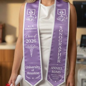 Puede incluir: Una mujer con un vestido blanco y una banda de graduación morada que dice "Sonographer" y "University of Houston". La banda tiene un símbolo médico y el texto "Clase de 2026". Sostiene un diploma enrollado atado con una cinta roja.