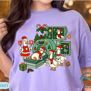 May include: A light purple t-shirt with a festive Christmas design. The illustration depicts cartoon ducks wearing Santa hats, some undergoing medical scans. The design includes candy canes, baubles, and a Christmas tree. The text "CC VIOLET" is at the bottom of the shirt.