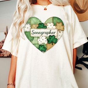 Könnte beinhalten: Cremefarbenes T-Shirt mit herzförmigem Design. Das Design zeigt einen Patchwork-Muster in Grün und Beige, Kleeblätter und das Wort "Sonographer" auf einem Banner. Das T-Shirt ist ein lässiger, bequemer Stil.