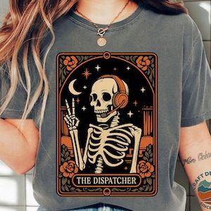 May include: Dark grey t-shirt with a graphic of a skeleton wearing headphones and giving a peace sign. The design includes a crescent moon, stars, and roses, with the text "THE DISPATCHER" in a decorative frame.