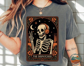 May include: Dark grey t-shirt featuring a graphic of a skeleton wearing headphones and giving a peace sign. The design includes a crescent moon, stars, and roses, with the text "THE DISPATCHER" in a decorative frame.