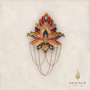 Puede incluir: Broche de cuentas con diseño de hoja, con colores rojo, dorado, negro y crema. Cadenas doradas cuelgan de la parte inferior. El broche está sobre un fondo color crema. El texto "sermeh atelier" está en la esquina inferior derecha.