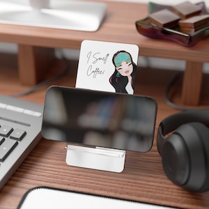 May include: A white and silver phone stand holding a black smartphone. A small white card with the text "I Smell Coffee!" and a cartoon illustration of a woman is behind the phone. Black headphones are on the right.