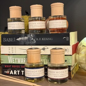 May include: Six glass bottles with wooden caps, each labelled with a different scent name, sit atop a stack of books. The labels include "Enchanted Timber," "Autumn Archives," "Cottage Cafe," "Bookshop Cafe," and "Amber Moon Ritual."