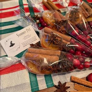 Puede incluir: Un conjunto de bolsitas para simmer pot con una tarjeta blanca que dice "Simmer Pot" con un logotipo de sombrero de bruja y el texto "Orange Slices, Cranberries, Cinnamon Stick, Star Anise & Cloves. Fill pot 3/4 full, bring water to boil, reduce heat and simmer for 1 hour. Do not add sugar or other sweeteners. Do not leave unattended."