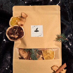 May include: A brown paper bag with a clear window showing dried orange slices, cinnamon sticks, and pine needles. The bag has a logo with a witch's hat and stars.