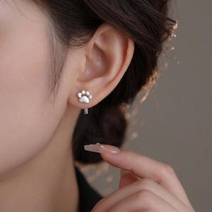 May include: A close-up of a person's ear with a silver paw print earring. The earring is a small, detailed representation of a paw, with the pads and claws clearly defined. The earring is attached to a small hoop. The background is blurred.