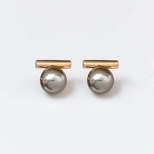 May include: A pair of stud earrings featuring a gold-colored bar above a gray, spherical bead. The earrings are displayed on a white background, showcasing their minimalist design and elegant aesthetic. The earrings are a jewelry item.