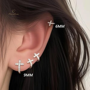 May include: Close-up of three silver cross earrings, one 6mm and two 9mm. The earrings are encrusted with small, sparkling stones. The earrings are displayed on an ear.