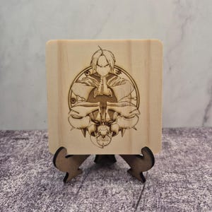 May include: A light-coloured wooden square with a detailed engraved image of an anime character with clasped hands, and a second character below. The square is displayed on a small wooden stand.