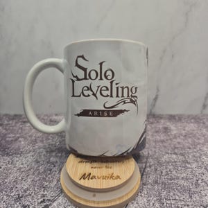Op de afbeelding: Witte keramische mok met de tekst "Solo Leveling: ARISE" in donkerbruin. De mok heeft een gebogen oor en staat op een houten onderzetter met de naam "Mavuika" en een inspirerende quote. Koffie- of theekop.