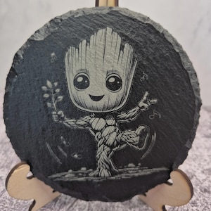 May include: Circular, dark grey slate coaster with an engraved image of Groot from Guardians of the Galaxy. The character is shown holding a sprig, with a smiling face and a tree-like form. The coaster is supported by a wooden stand.