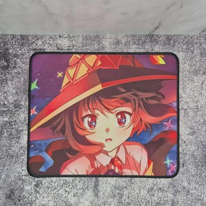 May include: Rectangular mouse mat featuring an anime-style illustration of a young female character with red hair and eyes, wearing a large red and yellow hat. The background is a starry night sky with colourful stars. The mouse mat has a black border.