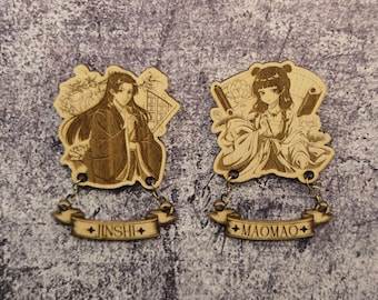 Pode incluir: Dois pingentes de madeira cortados a laser com ilustrações detalhadas de personagens em estilo anime. Cada pingente tem uma faixa com os nomes "JINSHI" e "MAOMAO". Os pingentes são conectados por pequenas correntes.