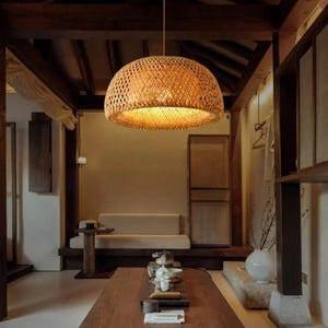 May include: A woven bamboo pendant light fixture hangs from the ceiling in a room with a wooden beam ceiling and light brown walls. The light fixture is round and has a natural brown color. The light fixture is illuminating the room with a warm, yellow light.