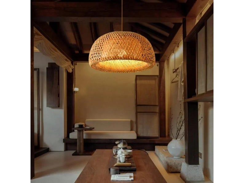 May include: A woven bamboo pendant light fixture hangs from the ceiling in a room with a wooden beam ceiling and light brown walls. The light fixture is round and has a natural brown color. The light fixture is illuminating the room with a warm, yellow light.