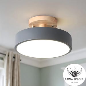 May include: A modern ceiling light with a grey cylindrical shade and a natural wood base. The light emits a bright, white glow and is mounted on a white ceiling. The fixture is a contemporary design element for home decor.