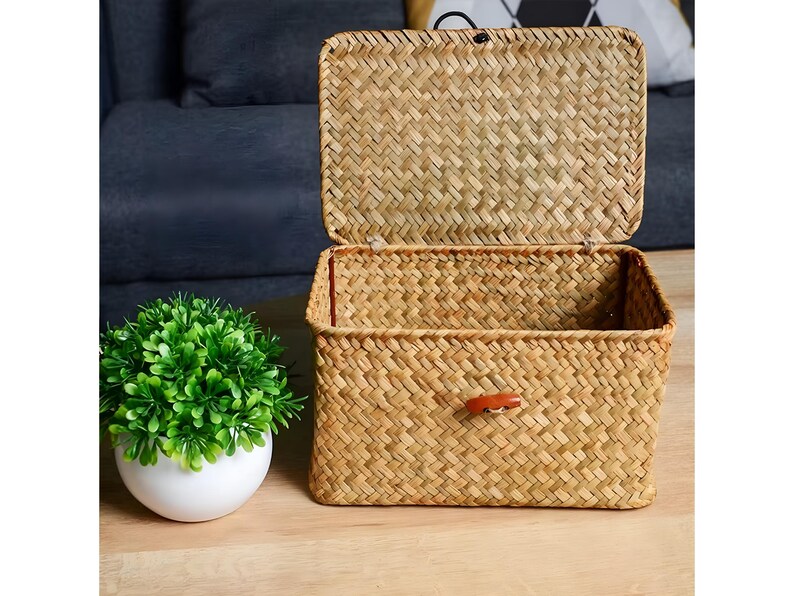 Puede incluir: Una cesta de almacenamiento rectangular tejida con tapa con bisagras abierta. La cesta est&aacute; hecha de material tejido de color natural y presenta una peque&ntilde;a leng&uuml;eta de cuero en la parte delantera. A la izquierda hay una peque&ntilde;a planta verde en una maceta blanca.