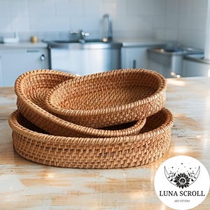 May include: Three woven rattan oval baskets of varying sizes are stacked on a light-colored wooden surface. The baskets are a warm, natural brown color and have a textured appearance. The background shows a kitchen setting.