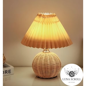 May include: A decorative table lamp with a woven rattan base and a pleated lampshade. The lampshade is a warm yellow color, and the base is a natural beige. The lamp is lit, casting a soft glow. The Luna Scroll Art Studio logo is visible.