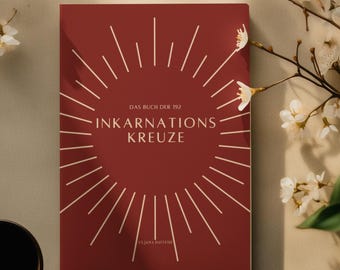 Puede incluir: Un libro granate titulado "DAS BUCH DER INKARNATIONS KREUZE" con un diseño de sol, sobre una superficie beige. Flores blancas y una taza negra están dispuestas alrededor del libro, creando una composición serena.