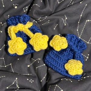 May include: A pair of blue knitted baby booties with yellow star accents. The booties are on a dark gray fabric with white constellations.