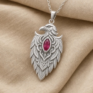 May include: A silver-toned metal pendant necklace featuring a detailed eagle head and wing design. The center of the pendant is set with a single, marquise-cut, deep red gemstone. The pendant hangs from a matching silver-toned metal link chain.