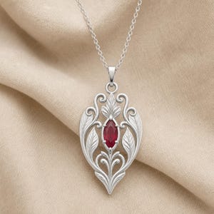May include: A silver-toned pendant necklace featuring a central marquise-cut deep red gemstone. The metal frame is crafted with intricate filigree scrollwork and leaf motifs, suspended from a delicate silver-toned chain link necklace.