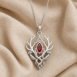May include: A silver-toned pendant necklace featuring a central marquise-cut deep red gemstone. The metalwork depicts two symmetrical deer antlers curving upward, with textured, vine-like branches intertwining at the base of the stone setting.