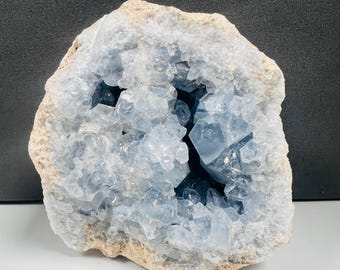 May include: A large, rough-edged celestite geode. The geode's exterior is a light tan color, while the interior reveals a cluster of pale blue crystals. The crystals are clustered together, creating a sparkling effect.