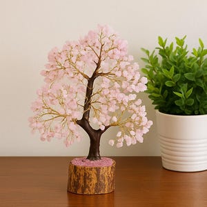 Könnte beinhalten: Eine dekorative Baumskulptur mit einem dunkelbraunen Stamm und Ästen, verziert mit rosa Edelsteinblättern. Der Baum steht auf einem Holzsockel mit rosa Akzenten. Eine grüne Topfpflanze befindet sich im Hintergrund.