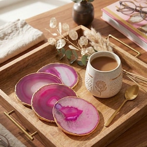 Peut inclure: Un plateau de service carré en bois avec des poignées en métal doré contenant quatre sous-verres circulaires en agate rose aux bords dorés, une tasse en céramique blanche tachetée avec un motif floral doré et une cuillère en métal doré.