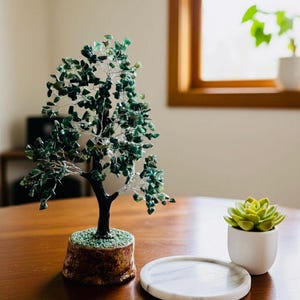 Può includere: Una scultura decorativa a forma di albero con foglie di gemme verdi e un tronco marrone scuro è appoggiata su una base di legno. Una piccola succulenta in un vaso bianco e un sottobicchiere di marmo sono anche sul tavolo di legno.